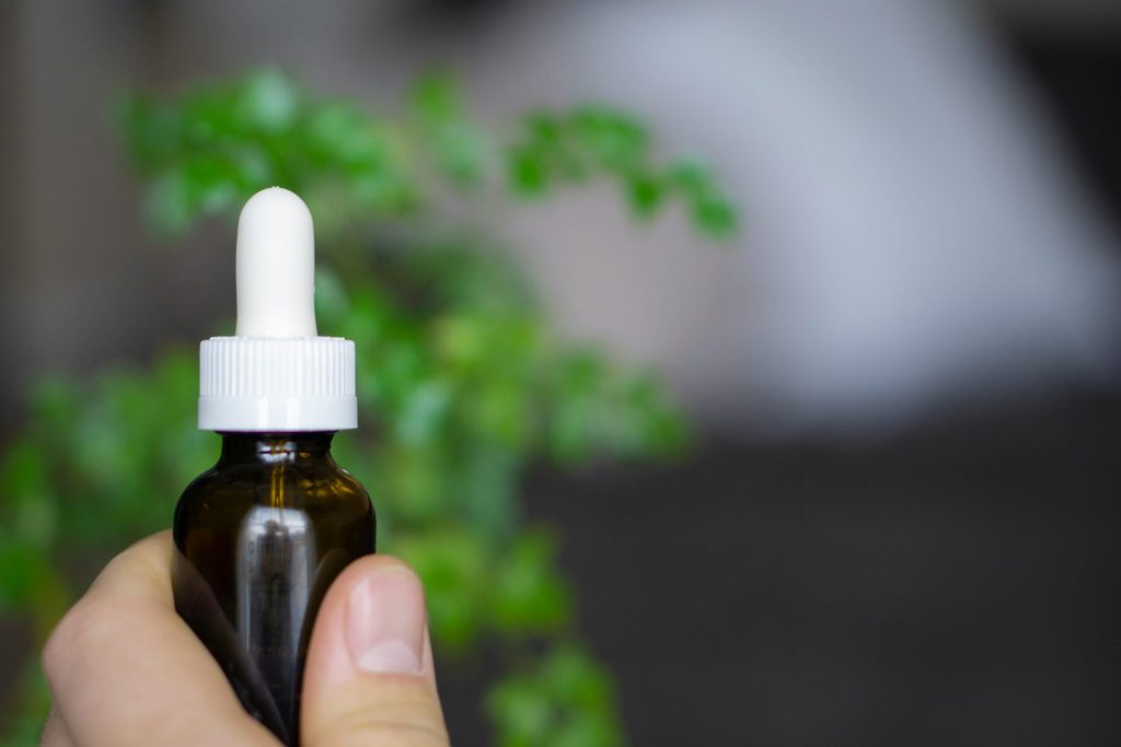 Eine Hand hält eine Tropfflasche mit CBD-Öl vor einem weichgezeichneten botanischen Hintergrund.